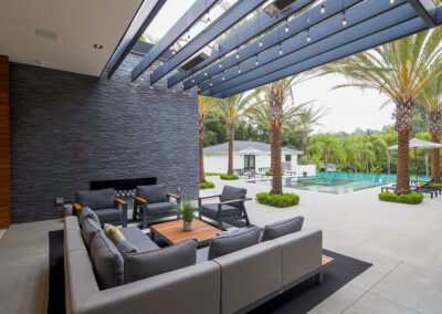 Modern outdoor patio with gray sectional sofas, a coffee table, and overhead string lights. Pool and palm trees visible in the background.