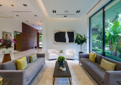 Modern living room with gray sofas, yellow pillows, a black-and-white artwork, and large windows overlooking greenery. Two white chairs are positioned near a tall indoor plant.