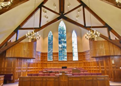 Interior of a church with wooden pews, exposed wooden beams, chandeliers, and a large stained glass window above the altar.