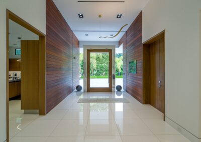 Modern entryway with wooden paneling and large glass doors. White tile floor, two spherical decor pieces, and ceiling light fixture. View of greenery through the doorway.