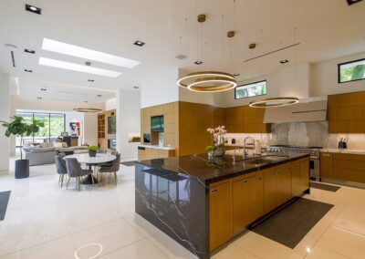 Modern kitchen and dining area with marble island, wooden cabinets, and circular pendant lights. Open layout extends to a living room with skylights and large windows.