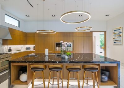 Modern kitchen with a large black marble island, wooden cabinets, and four barstools. Circular pendant lights hang above the island, and there's a stainless steel oven in the background.