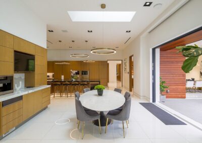 Modern kitchen with wooden cabinetry, a round marble dining table, gray chairs, and circular ceiling lights. Large open sliding doors lead to an outdoor area.