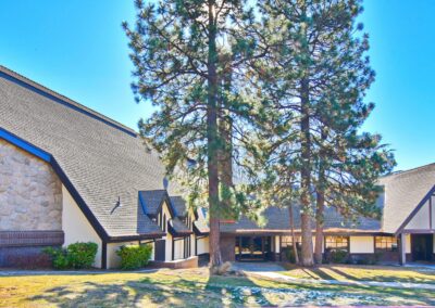 A-frame church building with stone and wood facade, large windows, and two tall pine trees in front, under a clear blue sky.