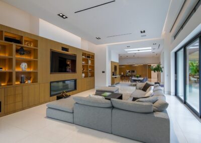 Modern living room with a large gray sectional sofa, wooden shelves, and a wall-mounted TV. Open layout leads to dining area with a large table. Natural light from sliding glass doors.