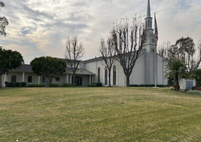 A large white church with a tall steeple and American flag, surrounded by leafless trees and a grassy lawn under a cloudy sky.