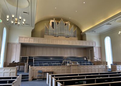 A church interior with rows of pews, a choir area, and a large pipe organ on an elevated platform. Chandelier lights hang from the ceiling.