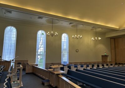 Interior of a church with empty pews, tall arched windows, and a podium. Light fixtures hang from the ceiling, and a clock is mounted on the wall.